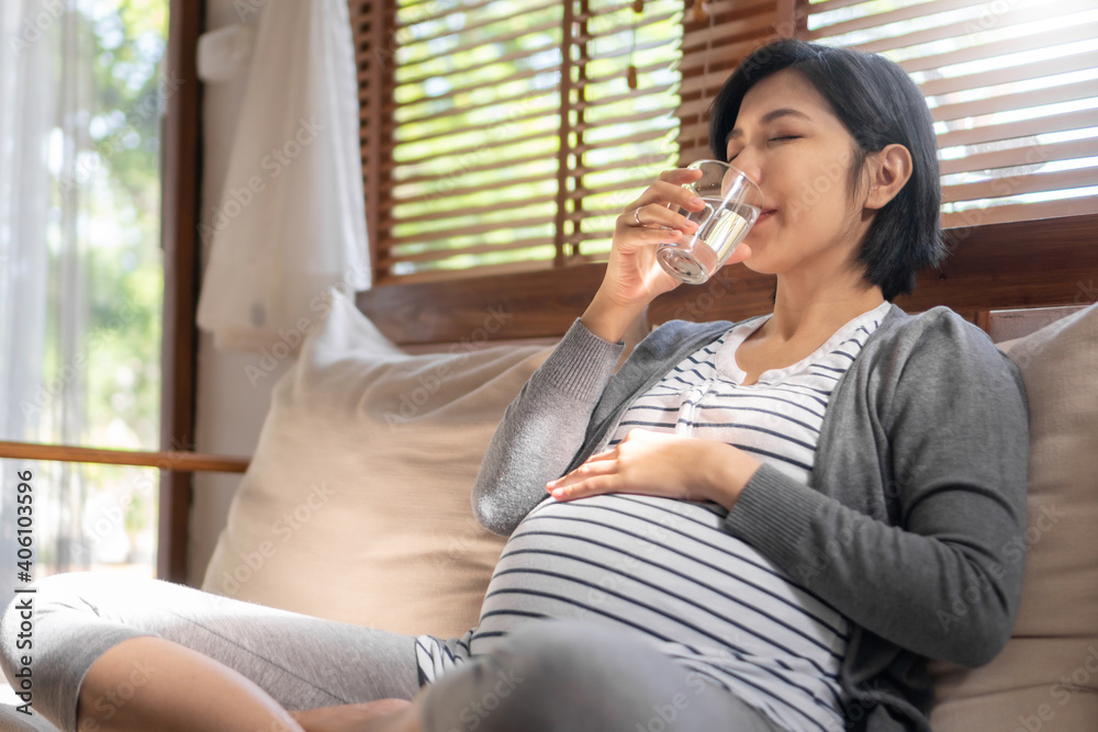 Young Asian pregnant woman sitting on sofa drinking pure fresh glass of water in the morning Young Asian pregnant woman sitting on sofa drinking pure fresh glass of water in the morning