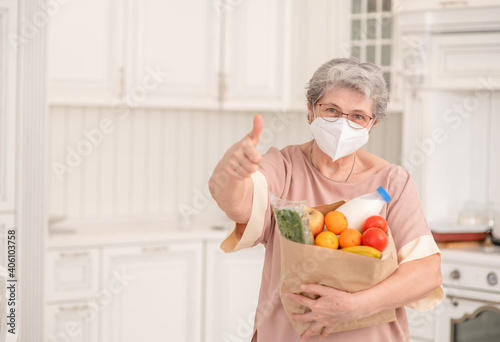 Senior woman wearing protective face mask holds bag with food and shows thumbs up gesture. Delivering food during quarantine Coronavirus (Covid-19) epidemic concept