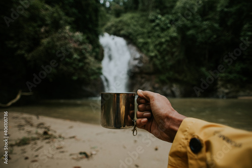 Close up shot of a man's hand holding coffee mug with waterfall in the background.Chilling during camping and hiking.