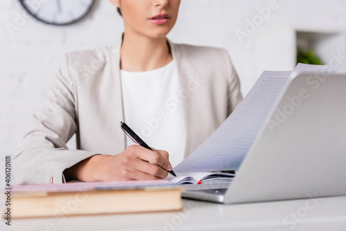 cropped view of translator writing in notebook while holding document near laptop, blurred foreground photo