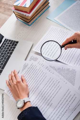 partial view of translator holding magnifier above documents near laptop and dictionaries of foreign languages photo