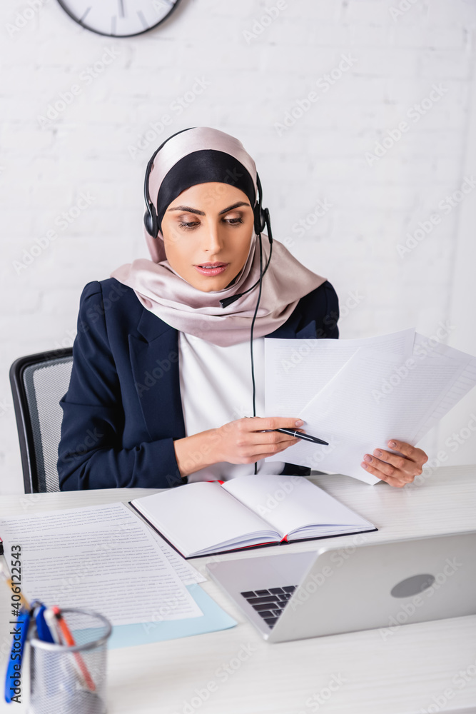 arabian interpreter in headset working with documents near blank notebook and laptop, blurred foreground arabian interpreter in headset working with documents near blank notebook and laptop, blurred foreground
