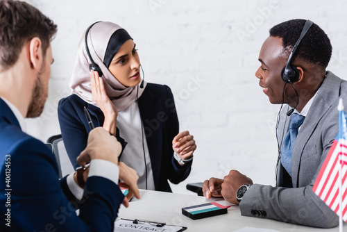 arabian and african american business partners in headsets near translator on blurred foreground photo