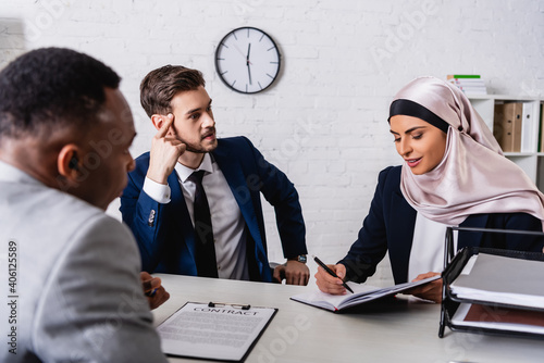 arabian businesswoman writing in notebook during negotiation with interracial business partners, blurred foreground