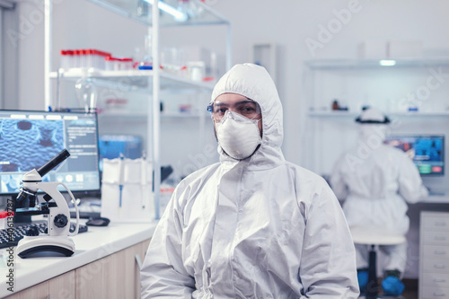 Chemist feeling tired from work for a long time in laboratory sitting at workplace. Overworked researcher dressed in protective suit against invection with coronavirus during global epidemic. photo