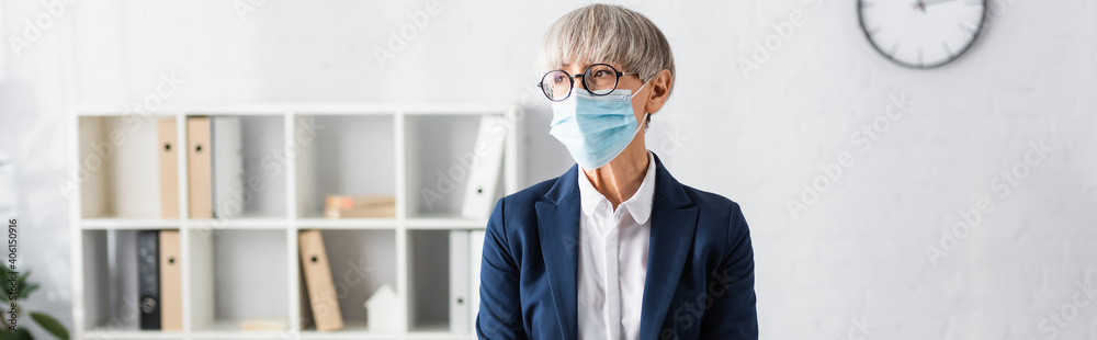 mature team leader in glasses and medical mask looking away in office, banner
