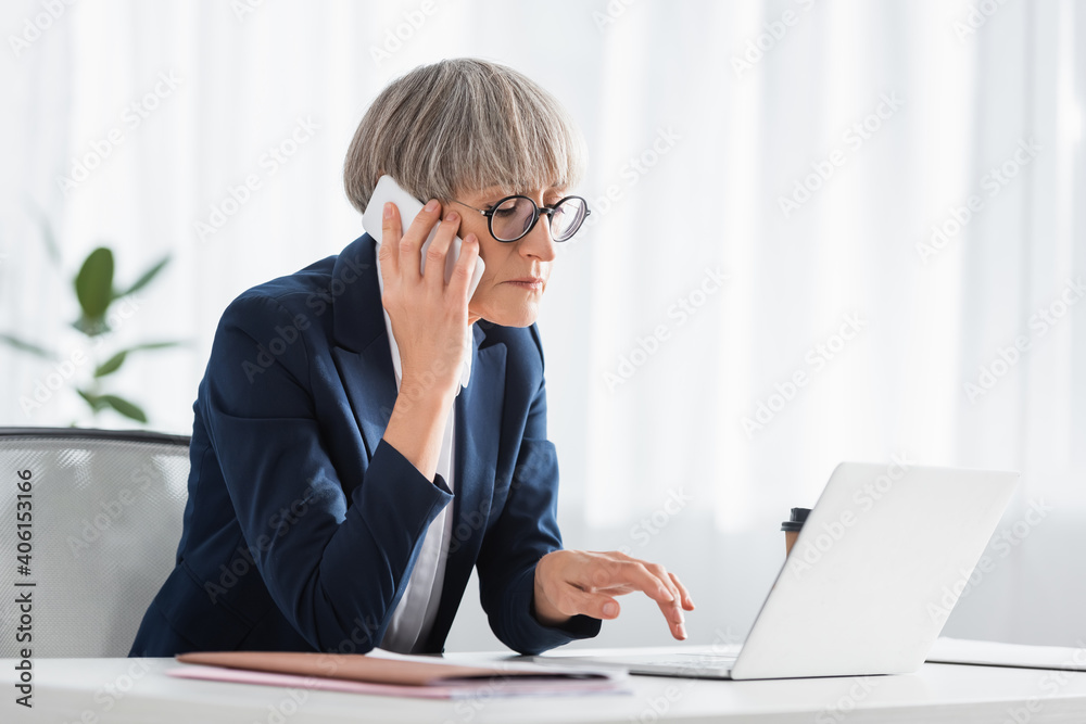 middle aged team leader in glasses talking on smartphone while using laptop middle aged team leader in glasses talking on smartphone while using laptop