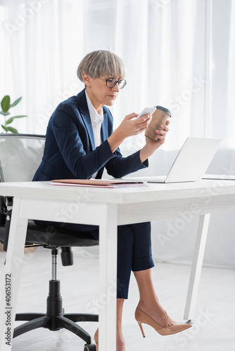 middle aged team leader in glasses holding coffee to go while using smartphone near laptop on desk © LIGHTFIELD STUDIOS