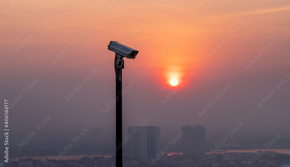Modern surveillance camera on cityscape and skyscraper background with sun shing bright before the sun was setting. The concept of surveillance evening time, copy space, Selective focus.