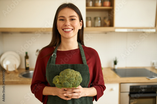 Cheerful young woman choosing healthy lifestyle, posing in kitchen with fresh raw broccoli in her hands, smiling at camera, going to bake vegetables in oven for dinner. Food, nutrition and dieting