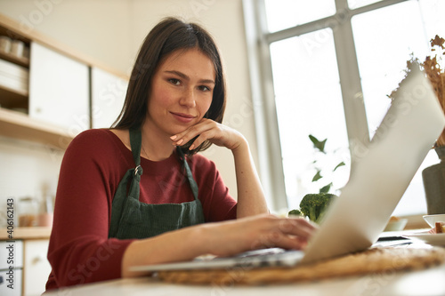 Portrait of cute young female freelancer working from home, sitting at kitchen table with portable computer, using high speed internet connection, making dinner at the same time, multitasking Free Download Portrait of cute young female freelancer working from home, sitting at kitchen table with portable computer, using high speed internet connection, making dinner at the same time, multitasking - GFXLoad
