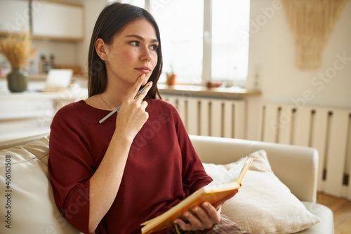 Thoughtful pensive young woman holding hand on her chin, having deep in thoughts look, thinking about future plans, making list of things to do in journal, sitting on couch in living room