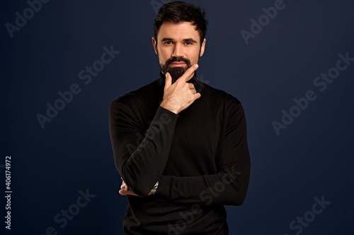 Stylish serious-looking young man with bristle in trendy high neck sweater, touching chin, rubbing beard and smirk as judging something, thinking or pondering checking out something interesting © Roman