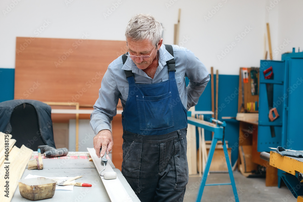 An elderly cabinetmaker in overalls and glasses paints a wooden board with a roller on a workbench in a carpentry shop. Non-staged photo while working with copy space