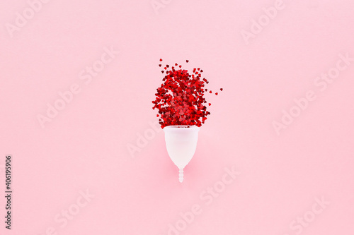 White menstrual Cup with red hearts symbolizing menstruation  on pink background. Alternative feminine hygiene product . Women health concept.
