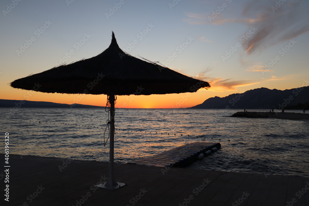 Sunset on the sea shore, silhouette of a beach umbrella against the background of an orangesky. Evening sky, silhouette of a reed beach umbrella on an empty beach on a warm autumn evening, Croatia