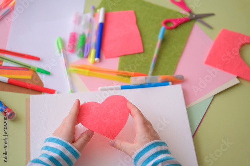 Сhild's boy's girl's hand holding heart cut out of felt. Children's creativity - drawings and crafts  for Valentines Day or Mother's Day or Father's Day. Top View.Colored paper, pens, scissors table photo