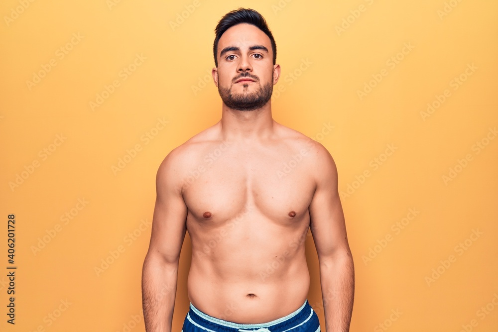 Young handsome man with beard wearing sleeveless t-shirt standing over yellow background with serious expression on face. Simple and natural looking at the camera.