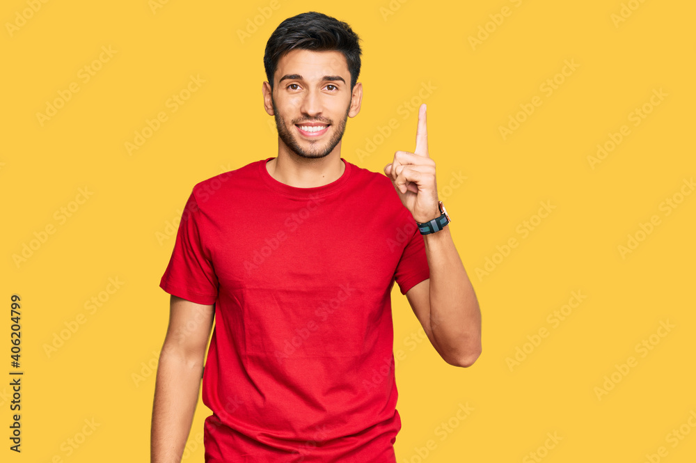 Young handsome man wearing casual red tshirt showing and pointing up with finger number one while smiling confident and happy. Young handsome man wearing casual red tshirt showing and pointing up with finger number one while smiling confident and happy.