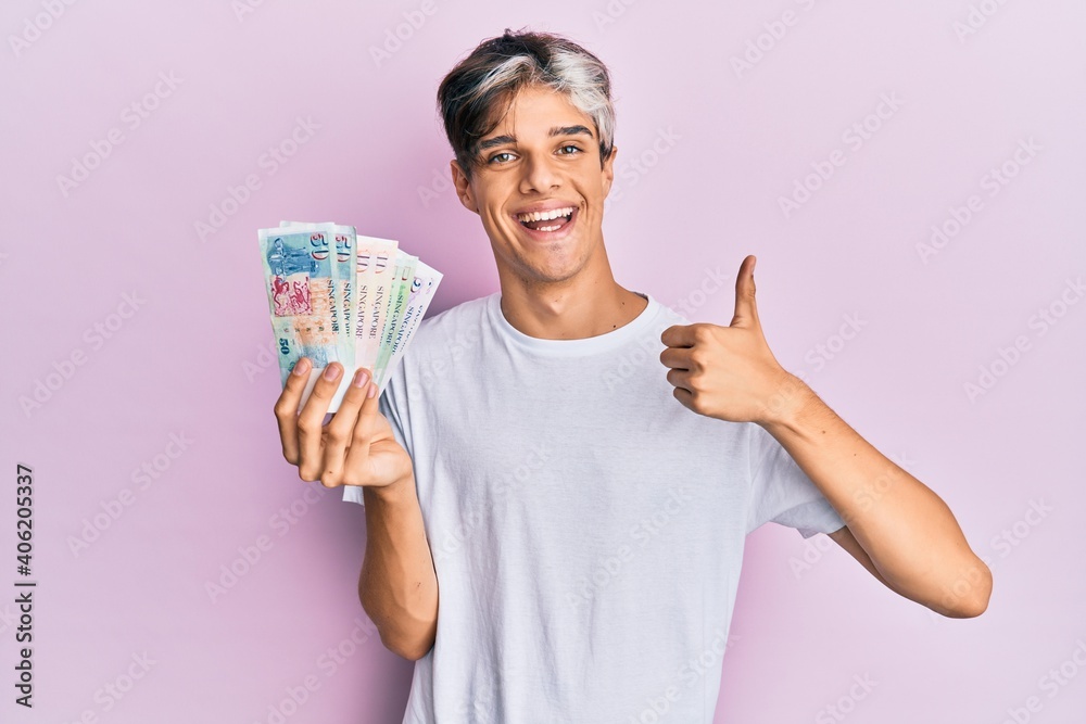 Young hispanic man holding singapore dollars banknotes smiling happy and positive, thumb up doing excellent and approval sign Young hispanic man holding singapore dollars banknotes smiling happy and positive, thumb up doing excellent and approval sign
