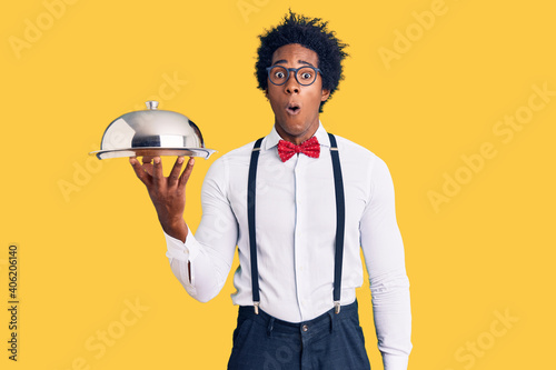 Handsome african american man with afro hair wearing waiter uniform holding silver tray scared and amazed with open mouth for surprise, disbelief face