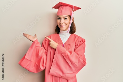 Young caucasian woman wearing graduation cap and ceremony robe amazed and smiling to the camera while presenting with hand and pointing with finger.