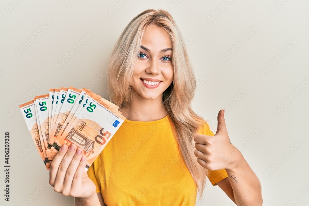 Beautiful caucasian blonde girl holding bunch of 50 euro banknotes smiling happy and positive, thumb up doing excellent and approval sign - obrazy, fototapety, plakaty 