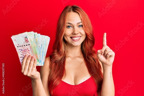 Young redhead woman holding hong kong dollars banknotes surprised with an idea or question pointing finger with happy face, number one