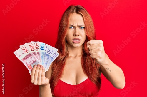Young beautiful redhead woman holding swedish krona banknotes annoyed and frustrated shouting with anger, yelling crazy with anger and hand raised