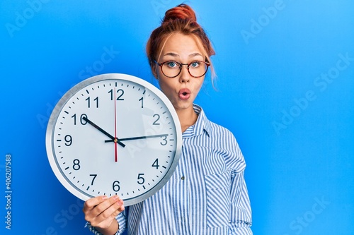 Young redhead woman holding big clock scared and amazed with open mouth for surprise, disbelief face
