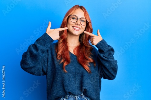 Young beautiful redhead woman wearing casual sweater and glasses over blue background smiling cheerful showing and pointing with fingers teeth and mouth. dental health concept. photo