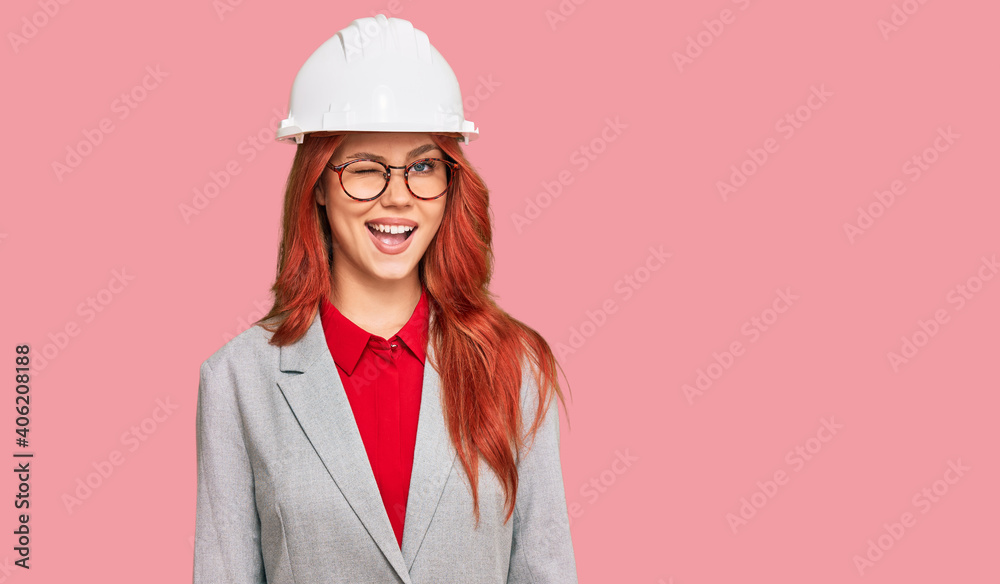 Young redhead woman wearing architect hardhat winking looking at the camera with sexy expression, cheerful and happy face.