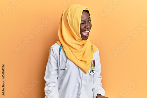 Beautiful african young woman wearing doctor uniform and hijab winking looking at the camera with sexy expression, cheerful and happy face. © Krakenimages.com