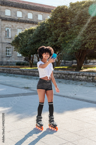 Young African american woman with afro hair takes a selfie with her mobile phone or makes a video call outside with inline roller skates to go skating training and playing sports in a sunny day