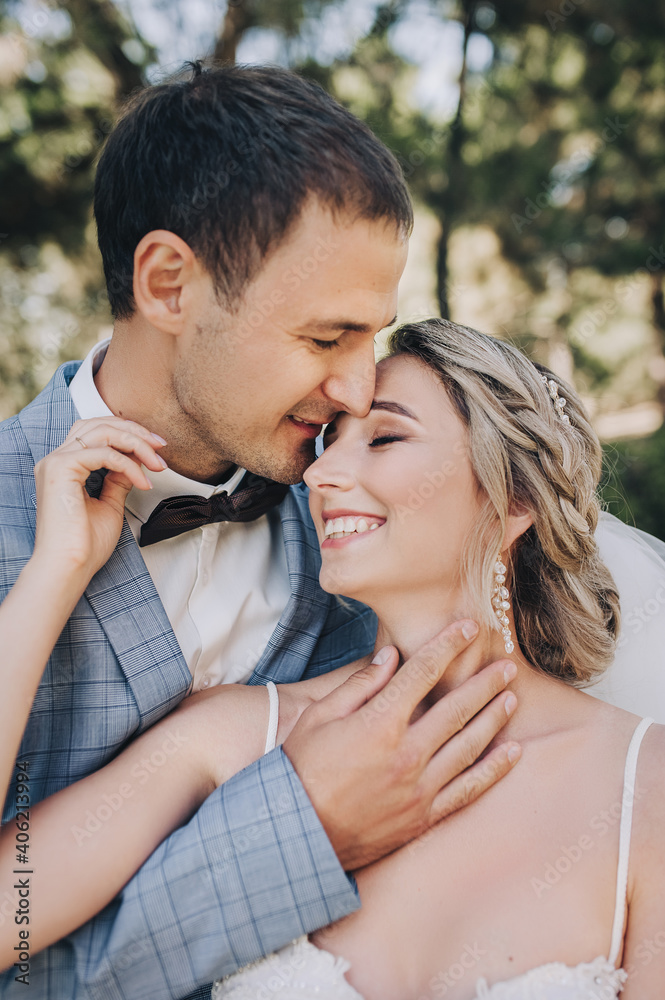 The groom in a gray checkered suit and a black bow tie and the blonde bride in a white lace dress tenderly hug in the park on the boulevard. Close-up wedding portrait of smiling newlyweds.