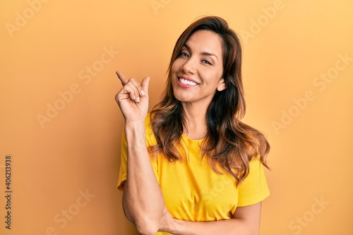 Young latin woman wearing casual clothes with a big smile on face, pointing with hand and finger to the side looking at the camera.