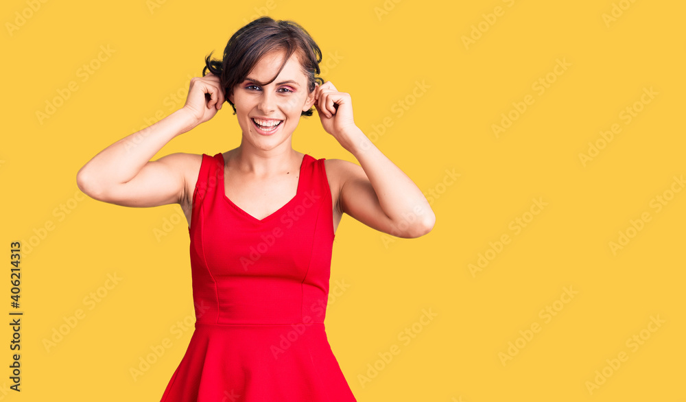 Beautiful young woman with short hair wearing casual style with sleeveless shirt smiling pulling ears with fingers, funny gesture. audition problem Beautiful young woman with short hair wearing casual style with sleeveless shirt smiling pulling ears with fingers, funny gesture. audition problem