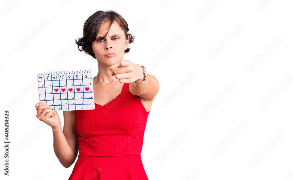 Beautiful young woman with short hair holding heart calendar pointing with finger to the camera and to you, confident gesture looking serious Beautiful young woman with short hair holding heart calendar pointing with finger to the camera and to you, confident gesture looking serious