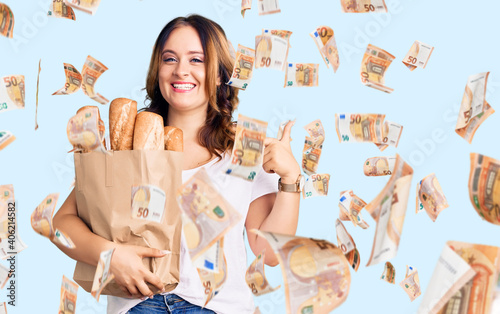 Young beautiful caucasian woman holding paper bag with bread pointing finger to one self smiling happy and proud