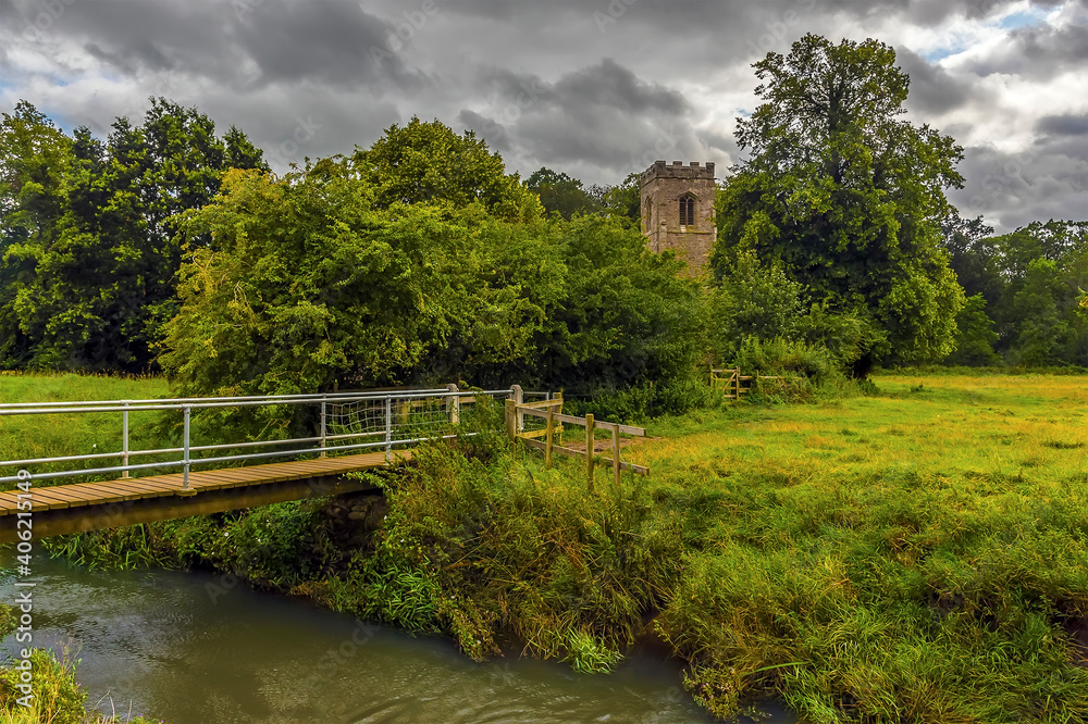 A rural path leads across the fields and streams towards a Norman church near Wistow, UK in summertime