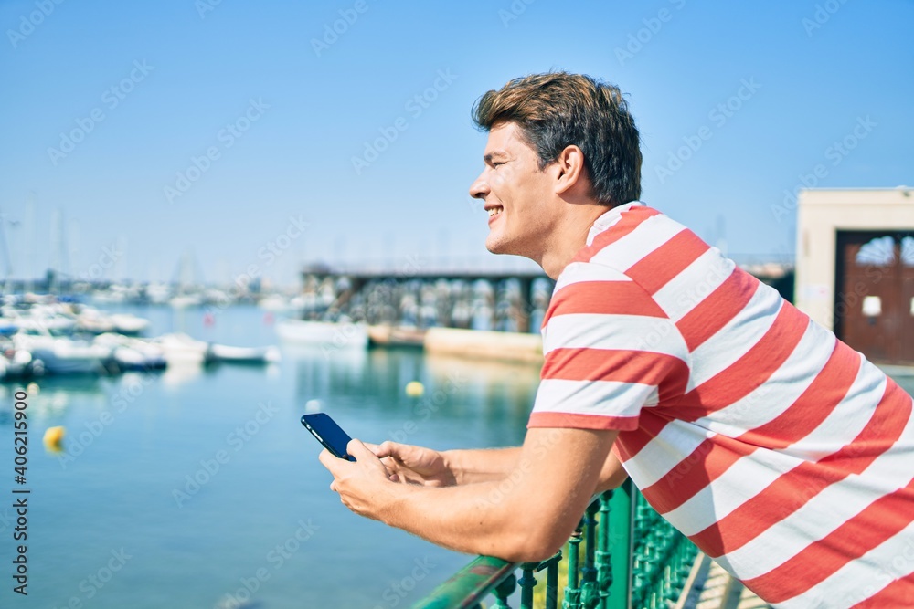 Young caucasian man smiling happy using smartphone leaning on the balustrade at the river.