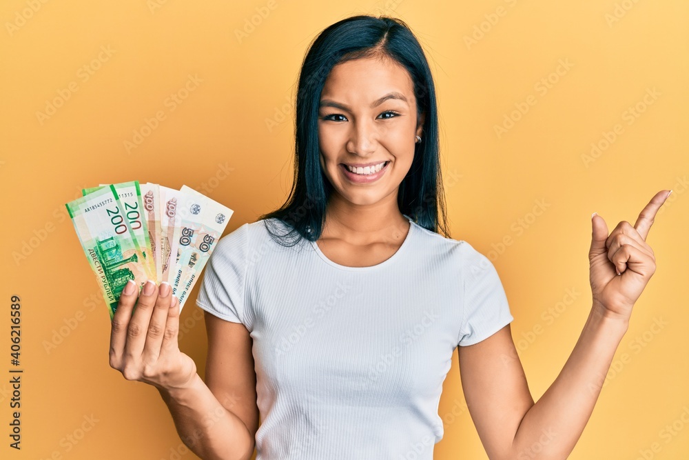 Beautiful hispanic woman holding russian ruble banknotes smiling happy pointing with hand and finger to the side
