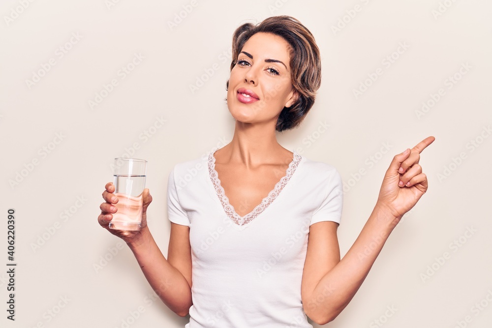 Young beautiful woman drinking glass of water smiling happy pointing with hand and finger to the side Young beautiful woman drinking glass of water smiling happy pointing with hand and finger to the side
