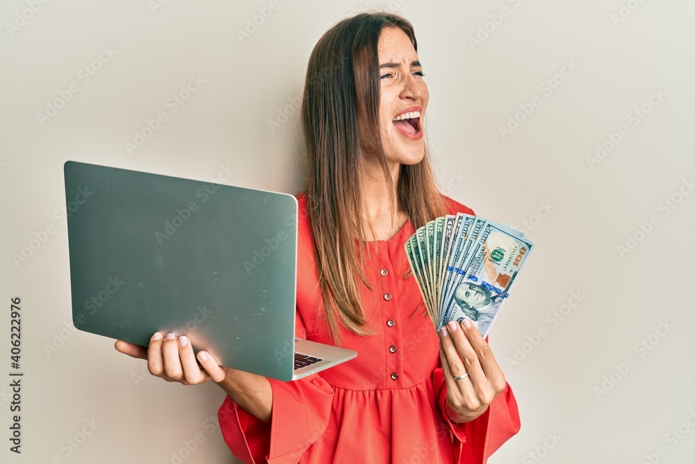 Young beautiful woman working at the office holding dollars angry and mad screaming frustrated and furious, shouting with anger. rage and aggressive concept. Young beautiful woman working at the office holding dollars angry and mad screaming frustrated and furious, shouting with anger. rage and aggressive concept.