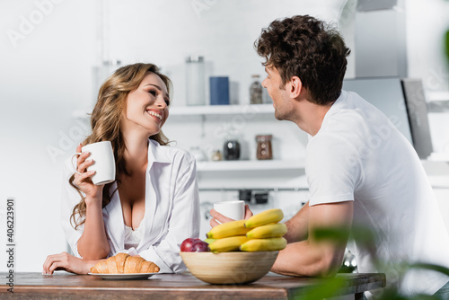 Smiling woman in shirt and bra holding cup near croissant, fruits and boyfriend on blurred foreground
