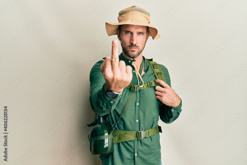 Handsome man with beard wearing explorer hat and backpack showing middle finger, impolite and rude fuck off expression Handsome man with beard wearing explorer hat and backpack showing middle finger, impolite and rude fuck off expression