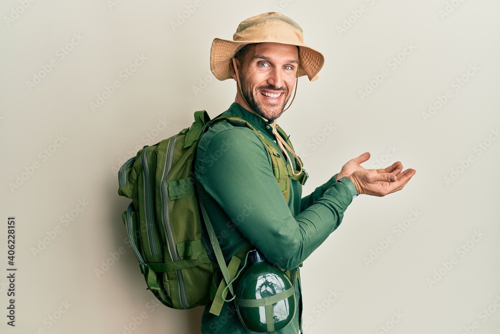 Handsome man with beard wearing explorer hat and backpack pointing aside with hands open palms showing copy space, presenting advertisement smiling excited happy Handsome man with beard wearing explorer hat and backpack pointing aside with hands open palms showing copy space, presenting advertisement smiling excited happy
