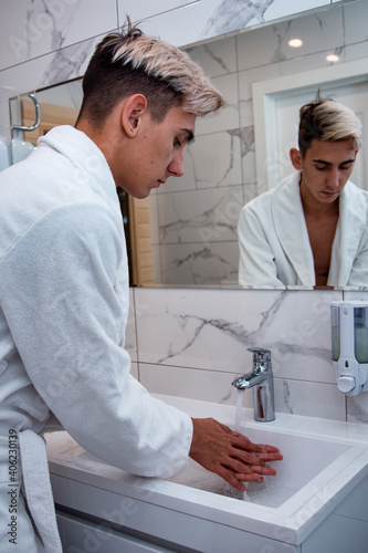 Morning procedures in the bathroom. A young man in a white coat stands at the mirror. Problem skin and care for it. Non-standard appearance.