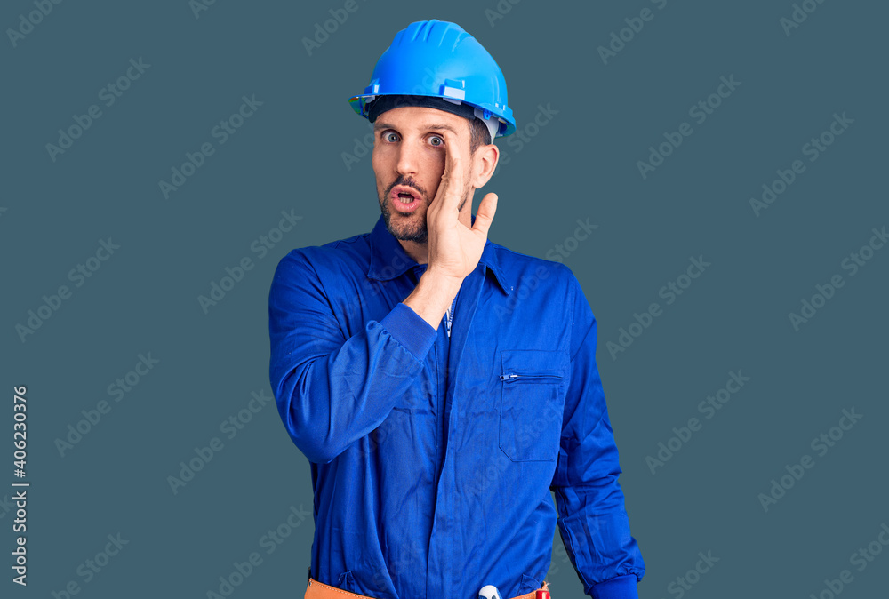 Young handsome man wearing worker uniform and hardhat hand on mouth telling secret rumor, whispering malicious talk conversation