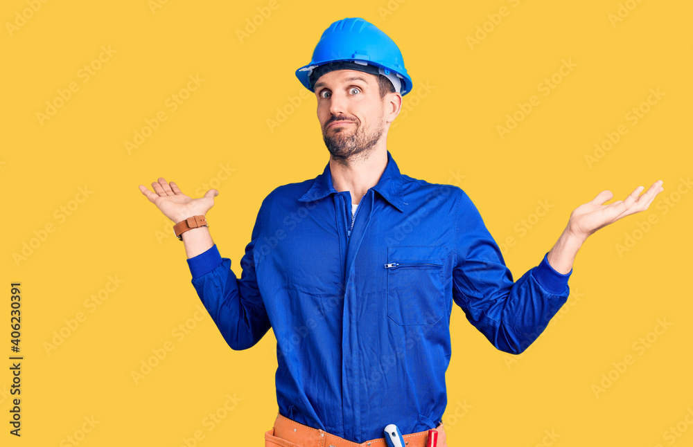 Young handsome man wearing worker uniform and hardhat covering ears with fingers with annoyed expression for the noise of loud music. deaf concept.